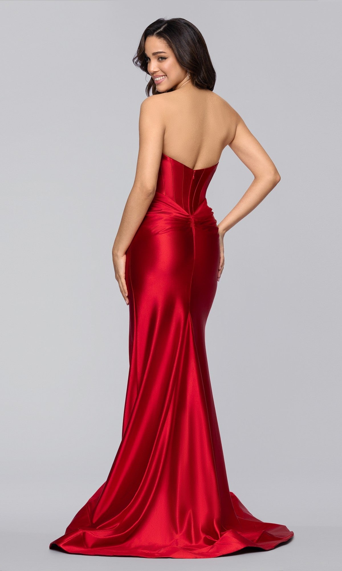 A woman models the Blondie Nites 5946BN, a strapless red satin long prom dress with a corset bodice, fitted skirt, and flowing train, posing with her back turned and glancing over her shoulder.